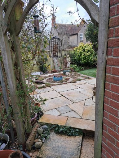 Stone pathway leading to a garden with plants and decorations, framed by an archway.